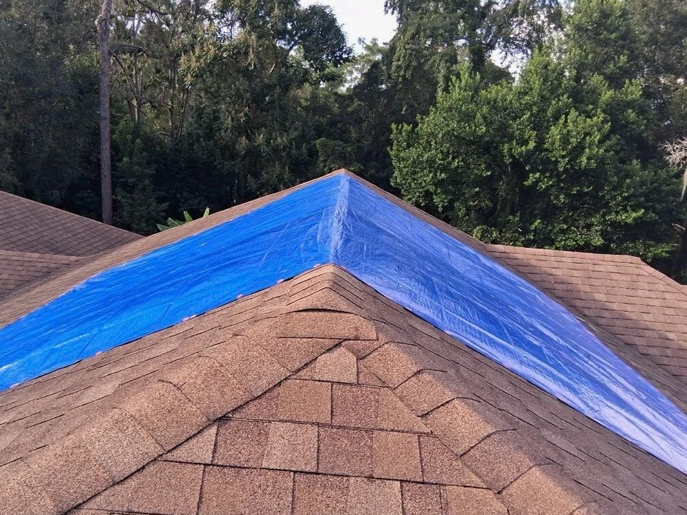 A blue emergency tarp fastened over both sides of a shingle roof ridge to keep the water out